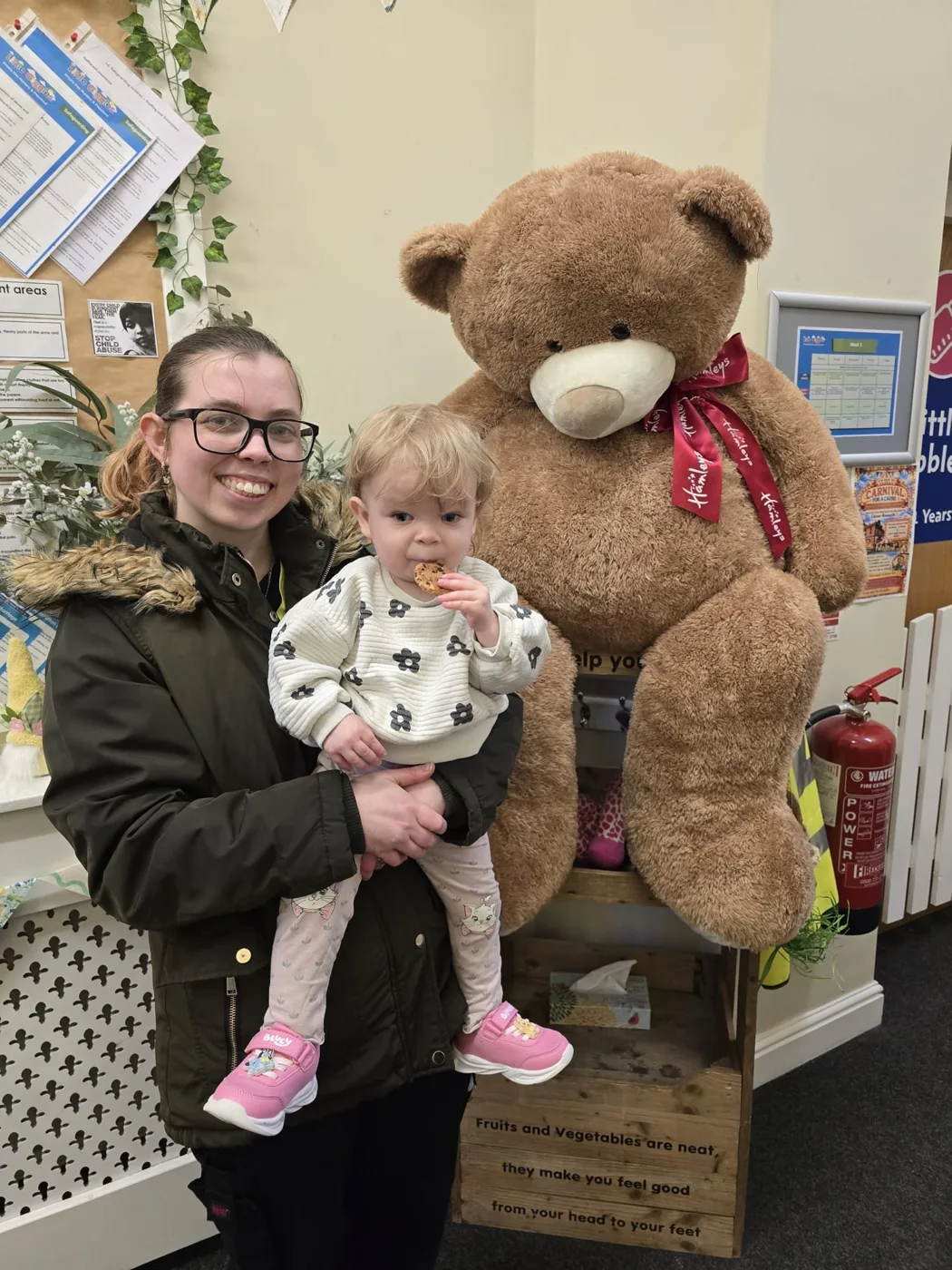 A woman holding a toddler eating a biscuit, standing beside the large Hamleys teddy bear in the nursery hallway