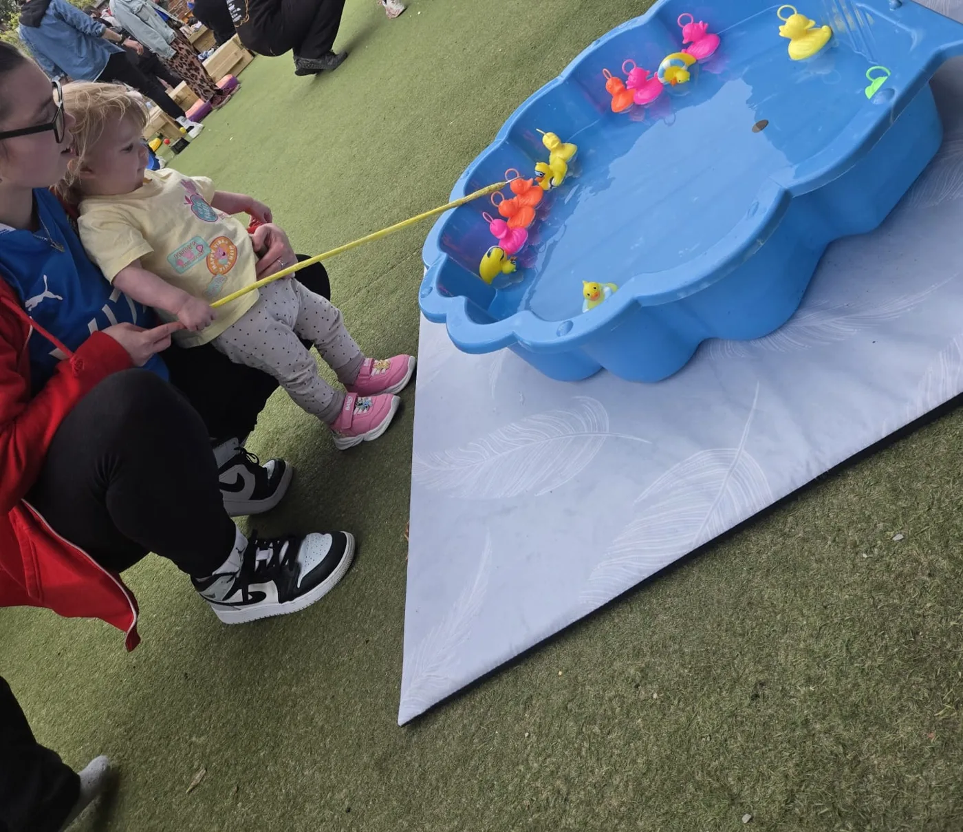 A toddler with a yellow fishing rod on an adult's lap, hooking plastic ducks from a blue paddling pool