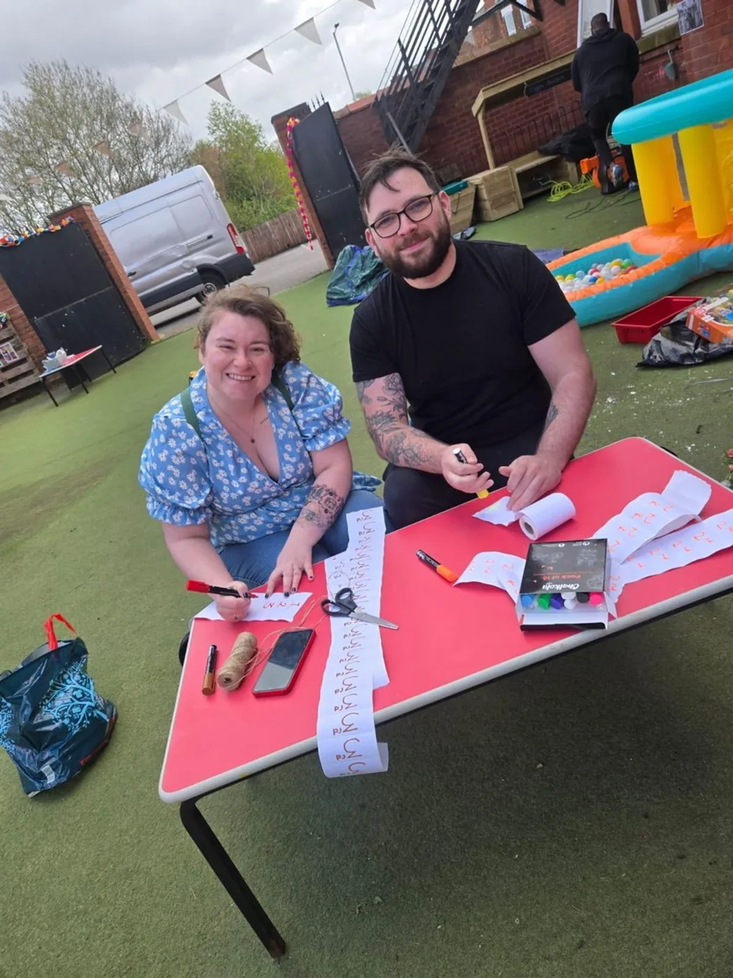 A woman and a man at a red table in the nursery garden writing out numbered tickets, with a bouncy castle behind them