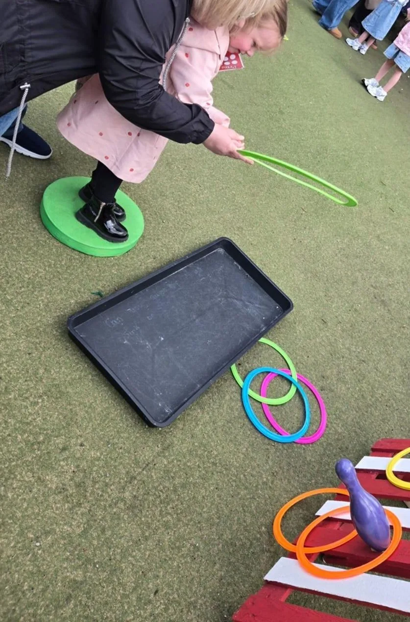 A child in a pink coat throwing a green hoop, with a skittle and hoops on a red and white pallet in the foreground