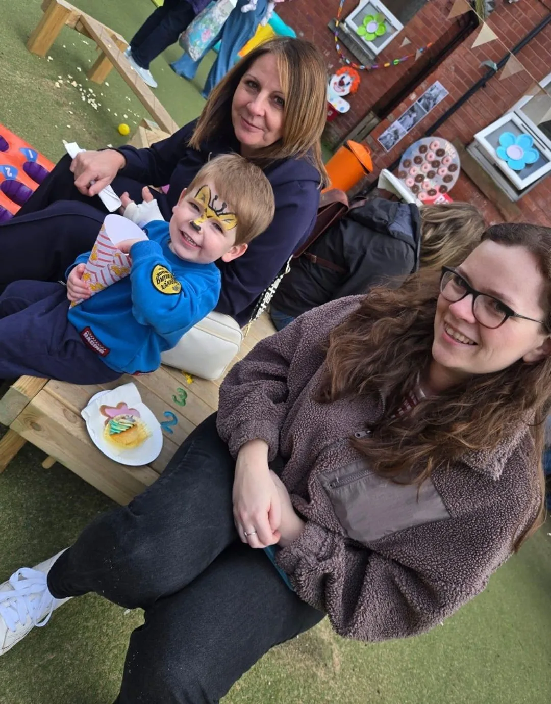 A boy with a gold face-painted crown and a cone of popcorn, sitting on a bench in the nursery garden with two women